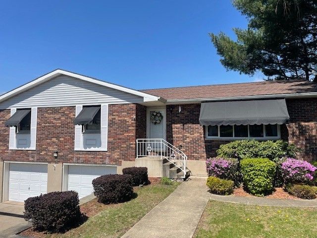 A split-level brick house with a two-car garage, white trim, grey awnings, and landscaping under a clear blue sky.