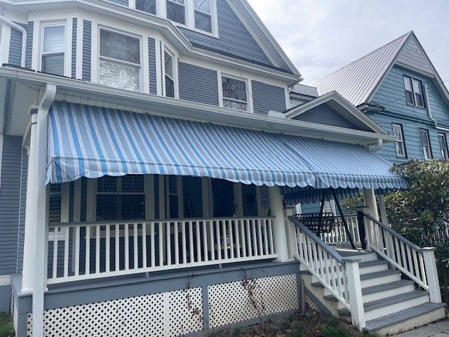 A blue and white striped awning covers the porch of a blue, multi-story house with a white railing and stairs.