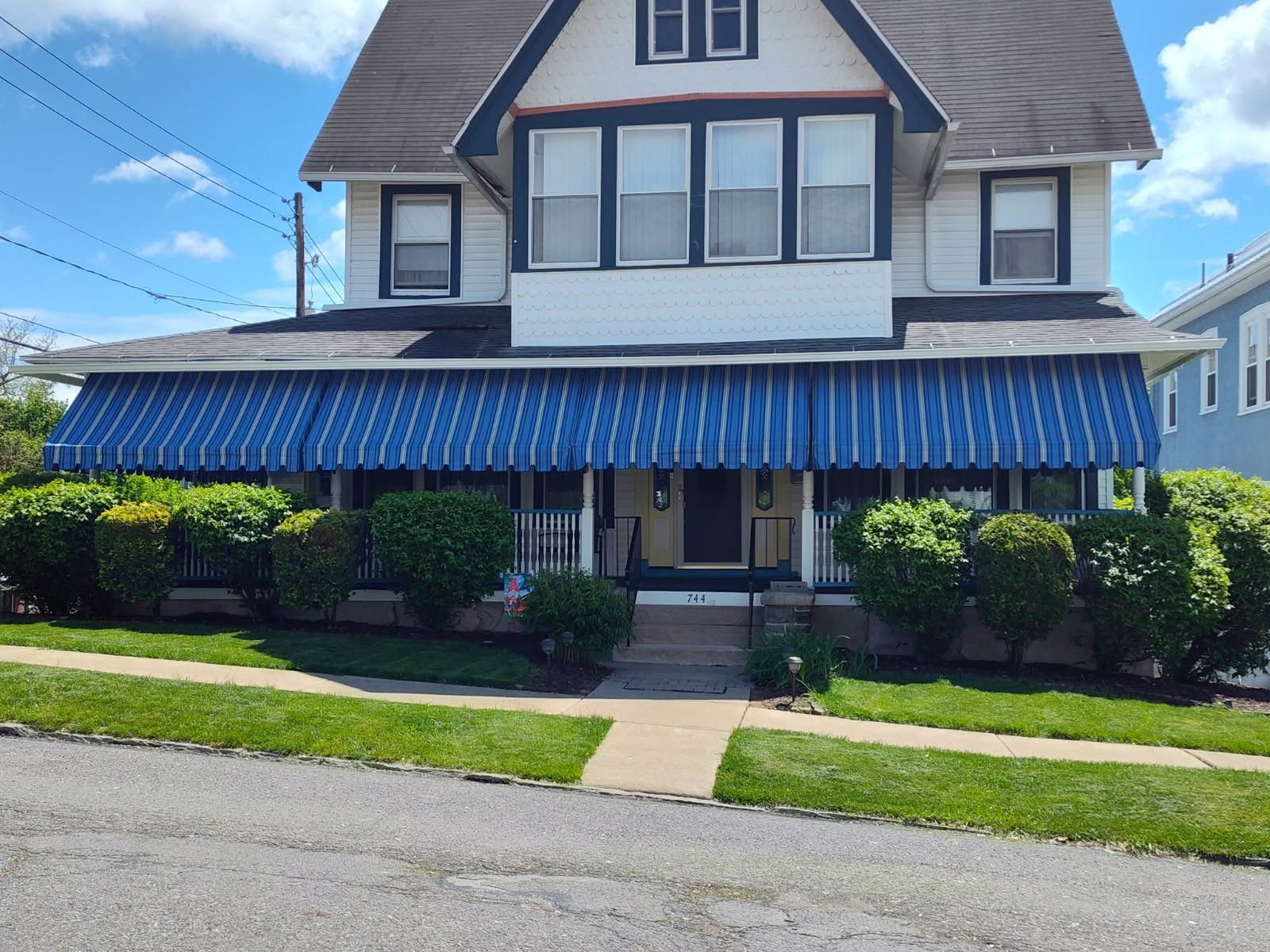 A two-story white house with a blue-and-white striped awning over the porch, surrounded by green shrubs and a lawn.