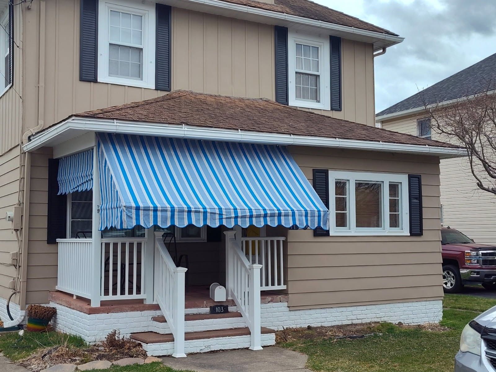 A tan two-story house features a white porch with a blue-and-white striped awning and a red truck parked in the driveway.