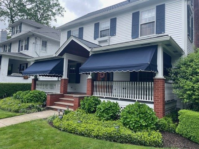 A white two-story house with blue shutters, a covered front porch featuring navy awnings, and landscaped front bushes.