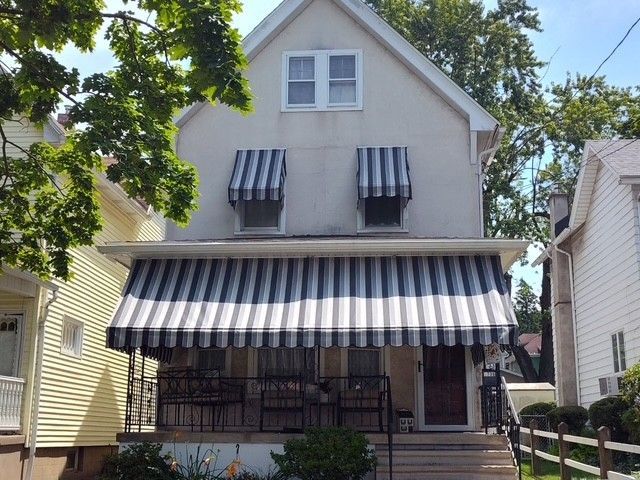 A two-story light-colored house with a front porch, covered by striped blue and white awnings over the windows and porch.