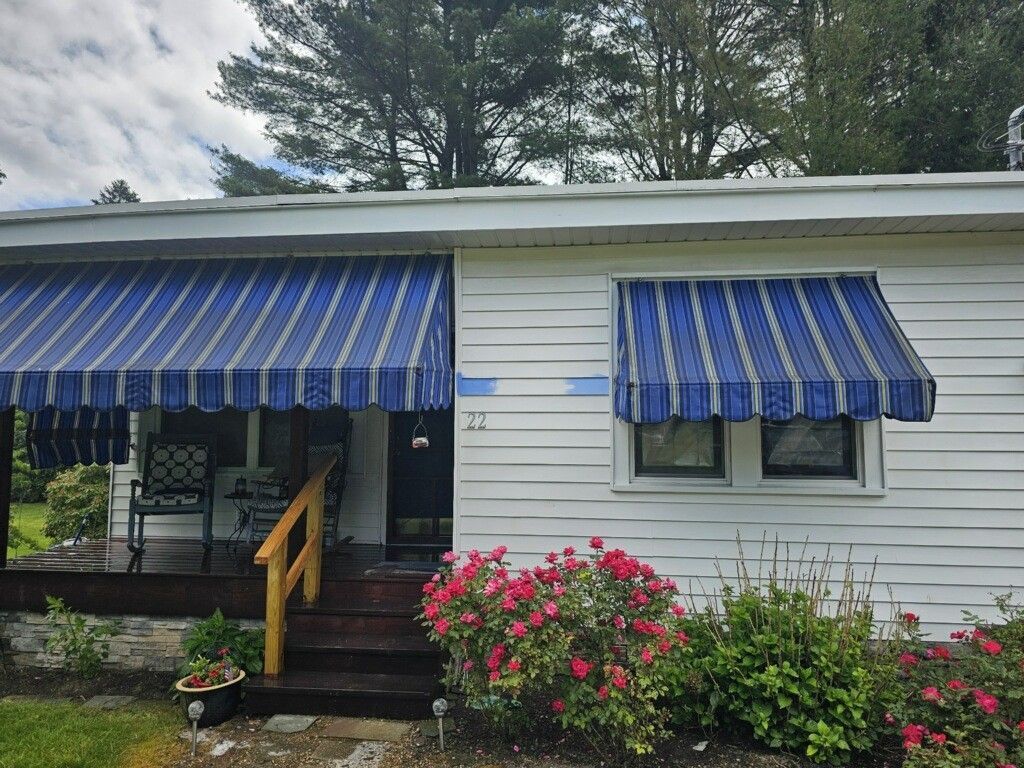 A single-story white house with blue and white striped awnings over the porch and window, and pink bushes in front.