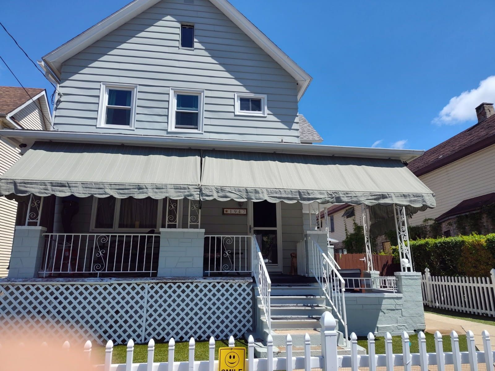 A two-story, light-gray house with a white front porch, lattice skirting, metal awning, and white picket fence.
