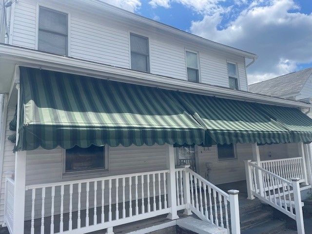 A two-story white house with a green and black striped awning covering the front porch and wooden railings.