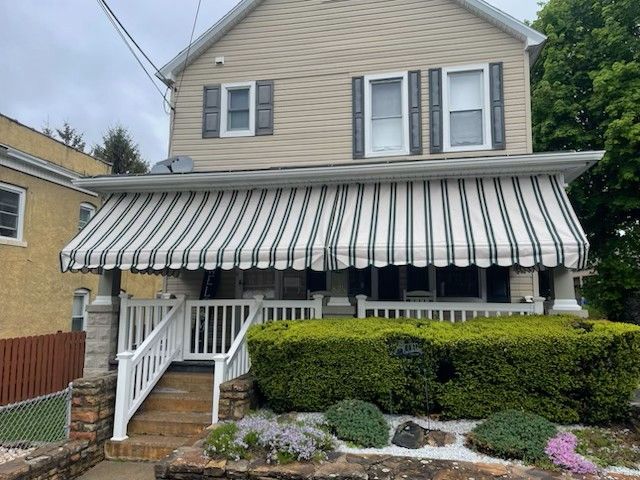 Two-story house with beige siding, a striped awning over the porch, white railings, and a landscaped front garden.