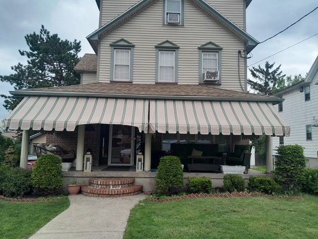 A two-story tan house with a wide, striped awning over the front porch, set behind a lawn and a concrete walkway.