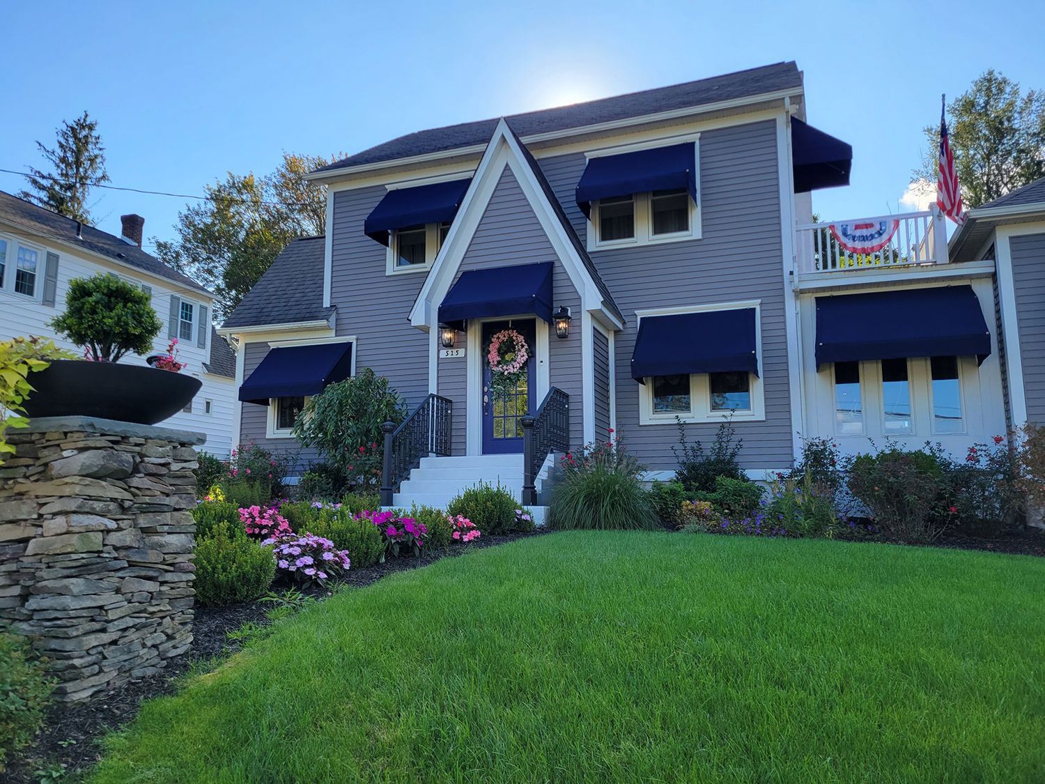 A gray two-story house with blue awnings, a stone pillar, and a neatly manicured lawn under a clear blue sky.