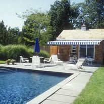 A swimming pool with white lounge chairs on a stone patio next to a cottage with a striped awning under a clear blue sky.