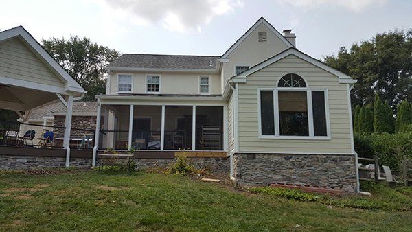 Rear exterior of a two-story house with a screened porch, light siding, and a stone foundation.