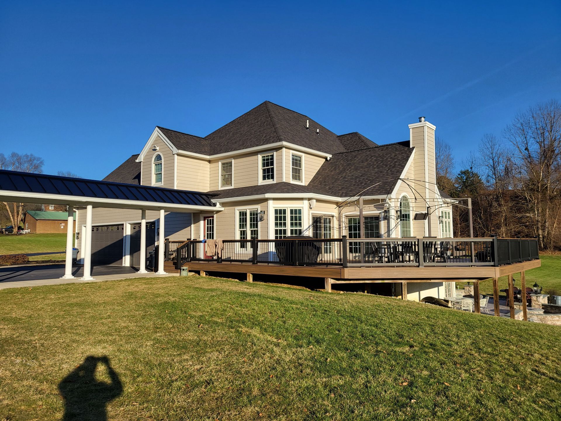 House with black roof, deck, carport, and chimney, on a grassy hill under a blue sky.