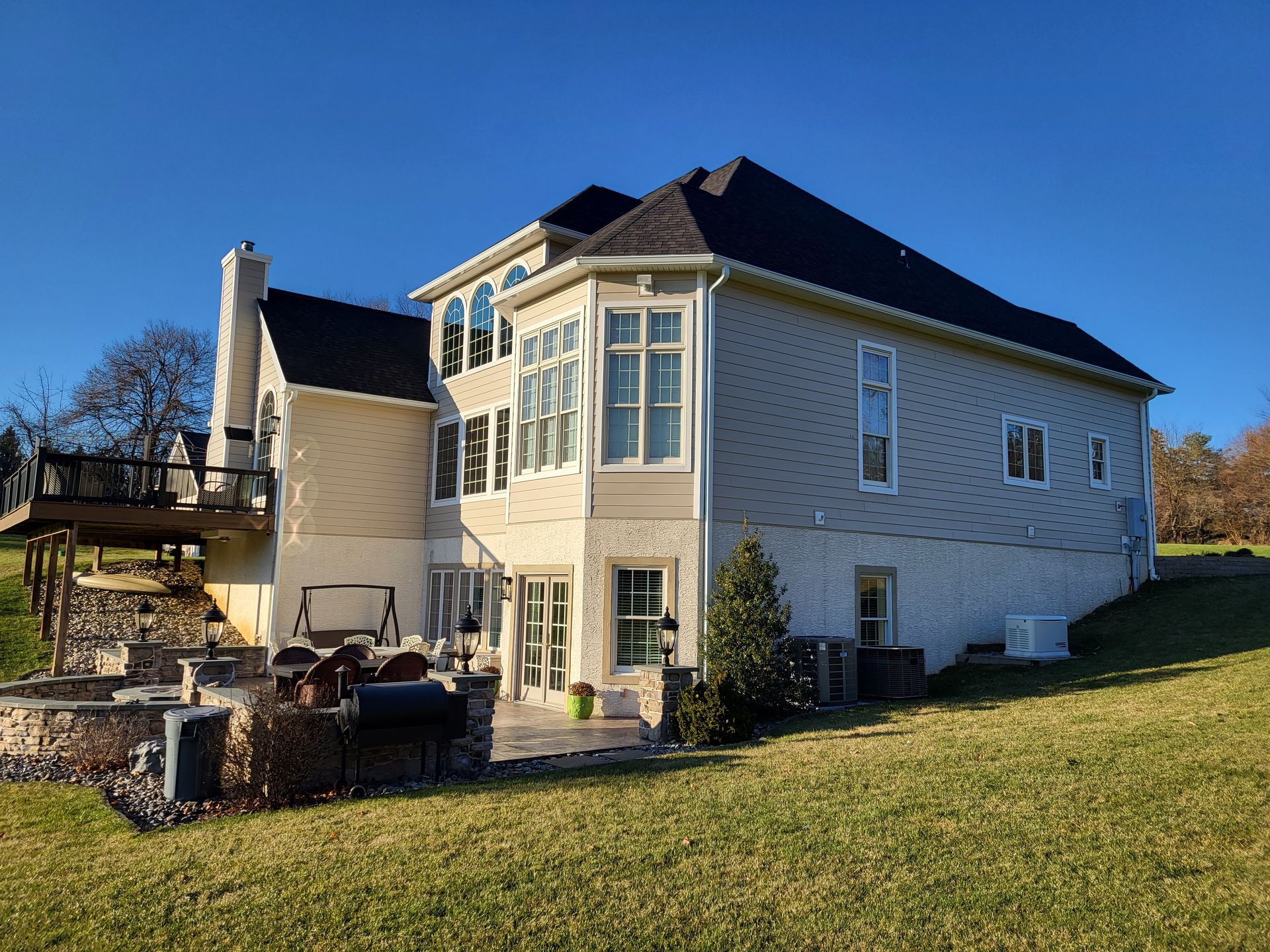 Large two-story house with beige siding, a black roof, and a bay window, set on a grassy hill under a blue sky.
