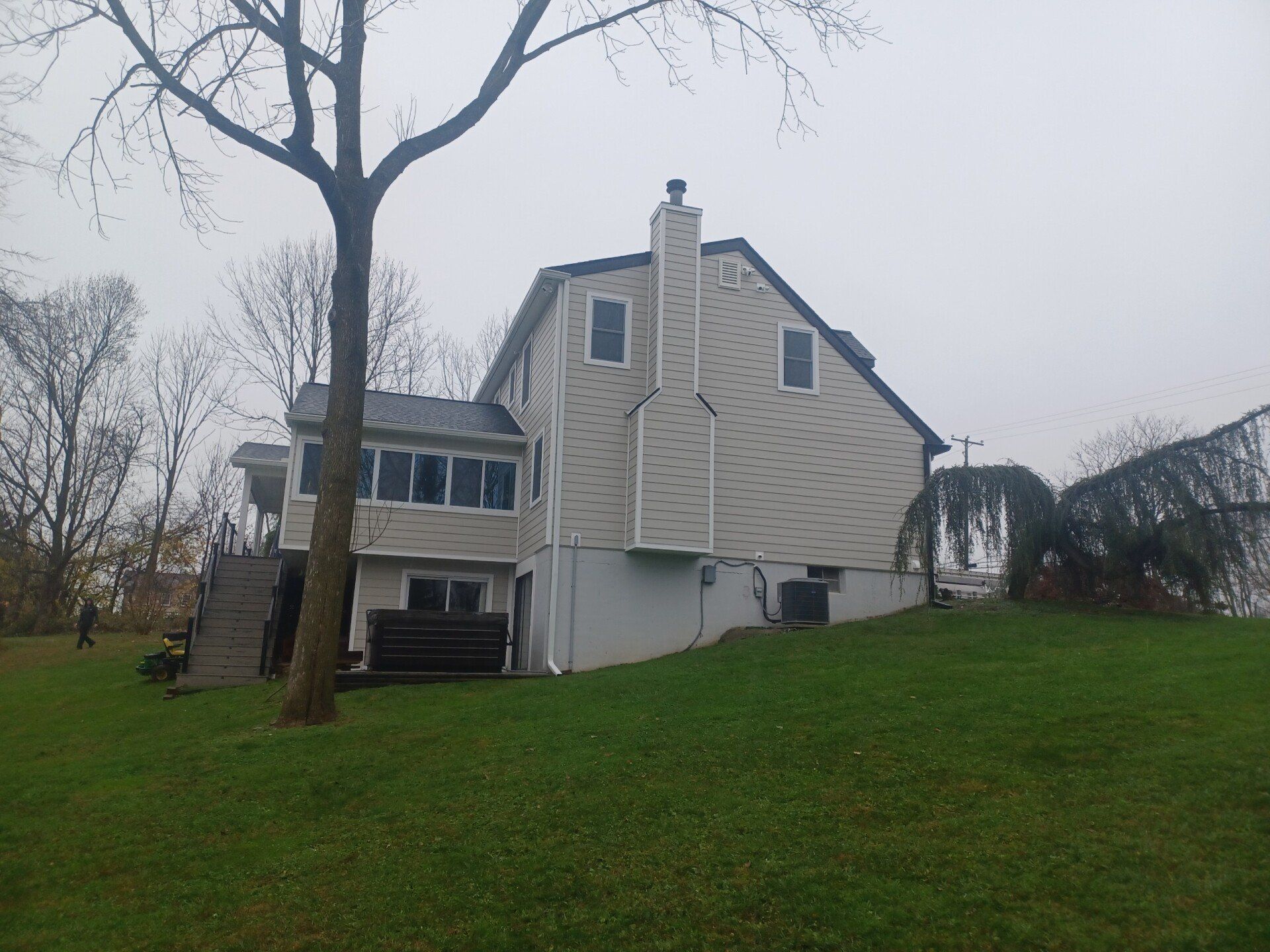 Back of a two-story house with a gray roof and light siding. Green grass slopes down from the house. Cloudy sky.