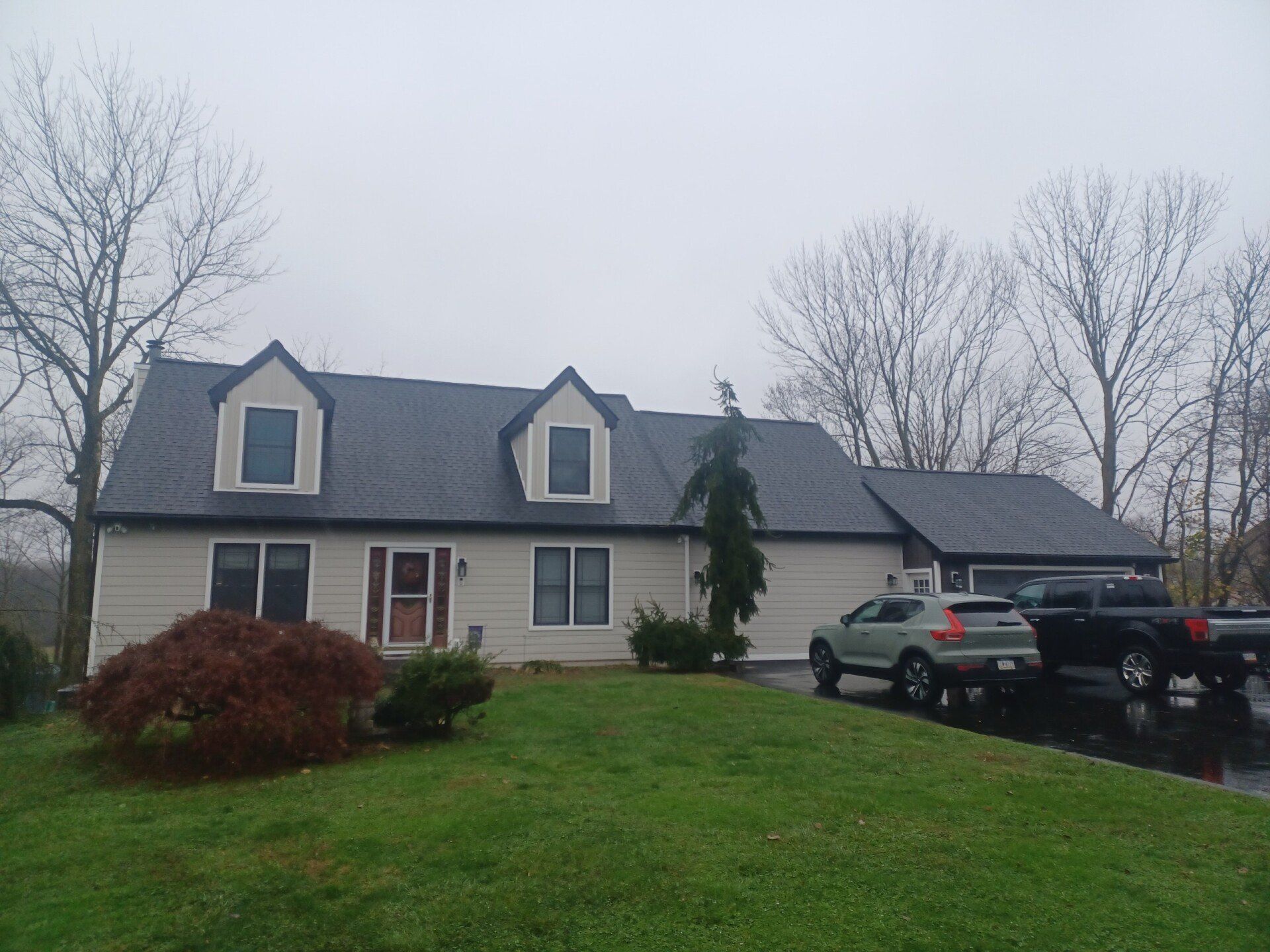 Beige house with dark roof and dormers; cars in driveway; overcast sky.