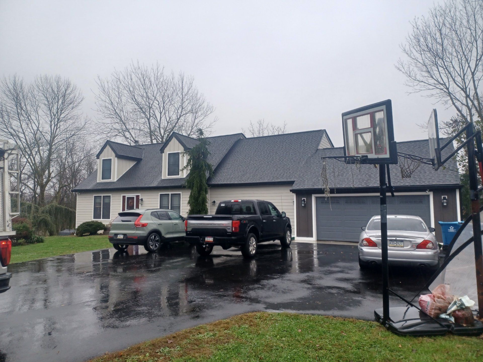 House with cars in the driveway on a rainy day; basketball hoop in the foreground.