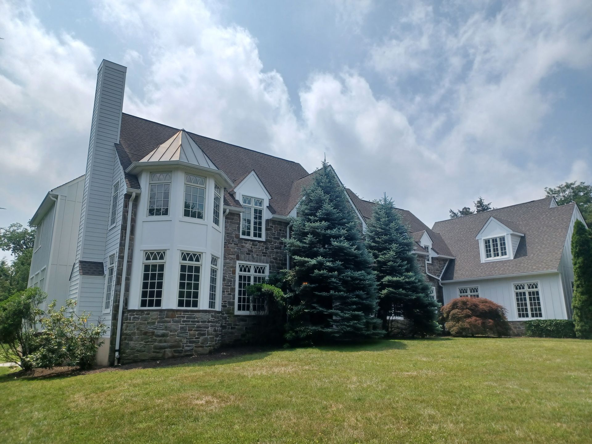 Large stone and white house with a brick chimney, evergreen trees, and a green lawn under a cloudy sky.