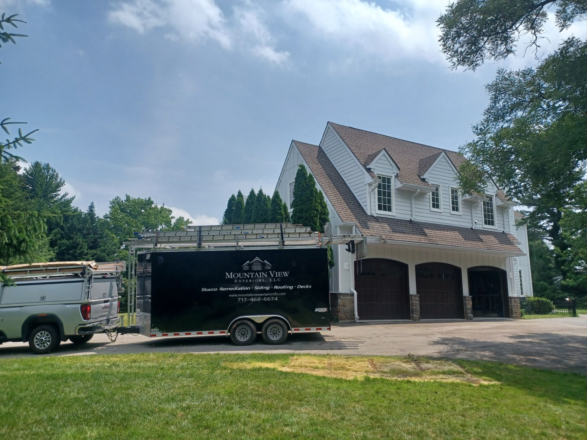 A white house with a brown roof has a trailer and truck in front of it on a sunny day.