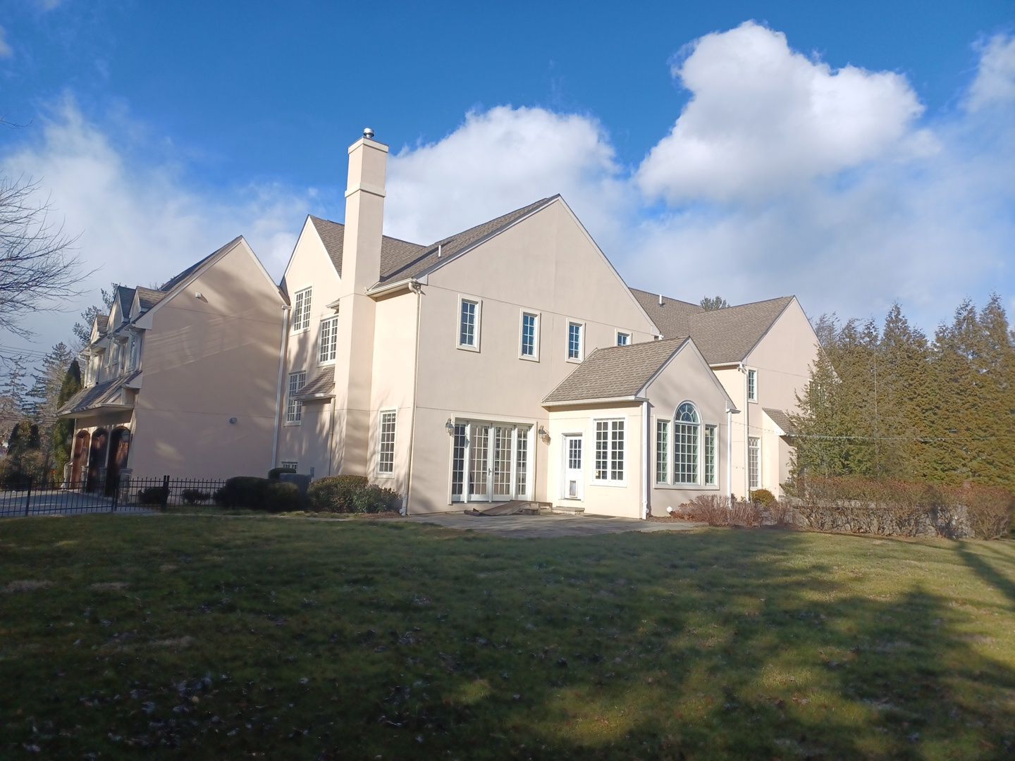 Beige two-story house with multiple rooflines, chimney, and windows, on a grassy lot with a blue sky and clouds.
