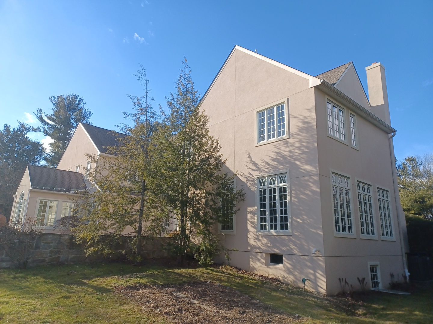 Beige house with white-framed windows, a chimney, and trees against a blue sky.