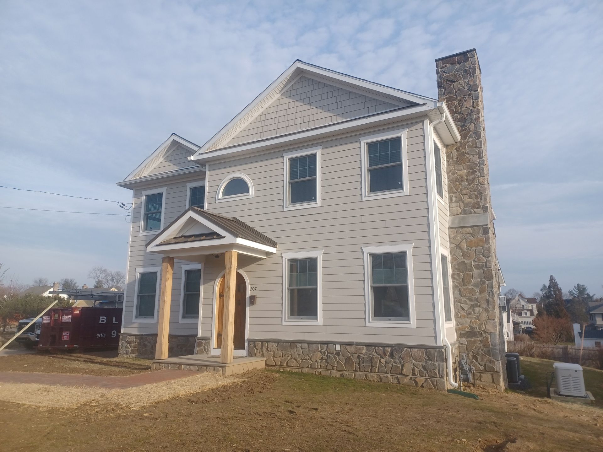 Two-story house with light gray siding and stone foundation. Tall stone chimney. Clear sky.