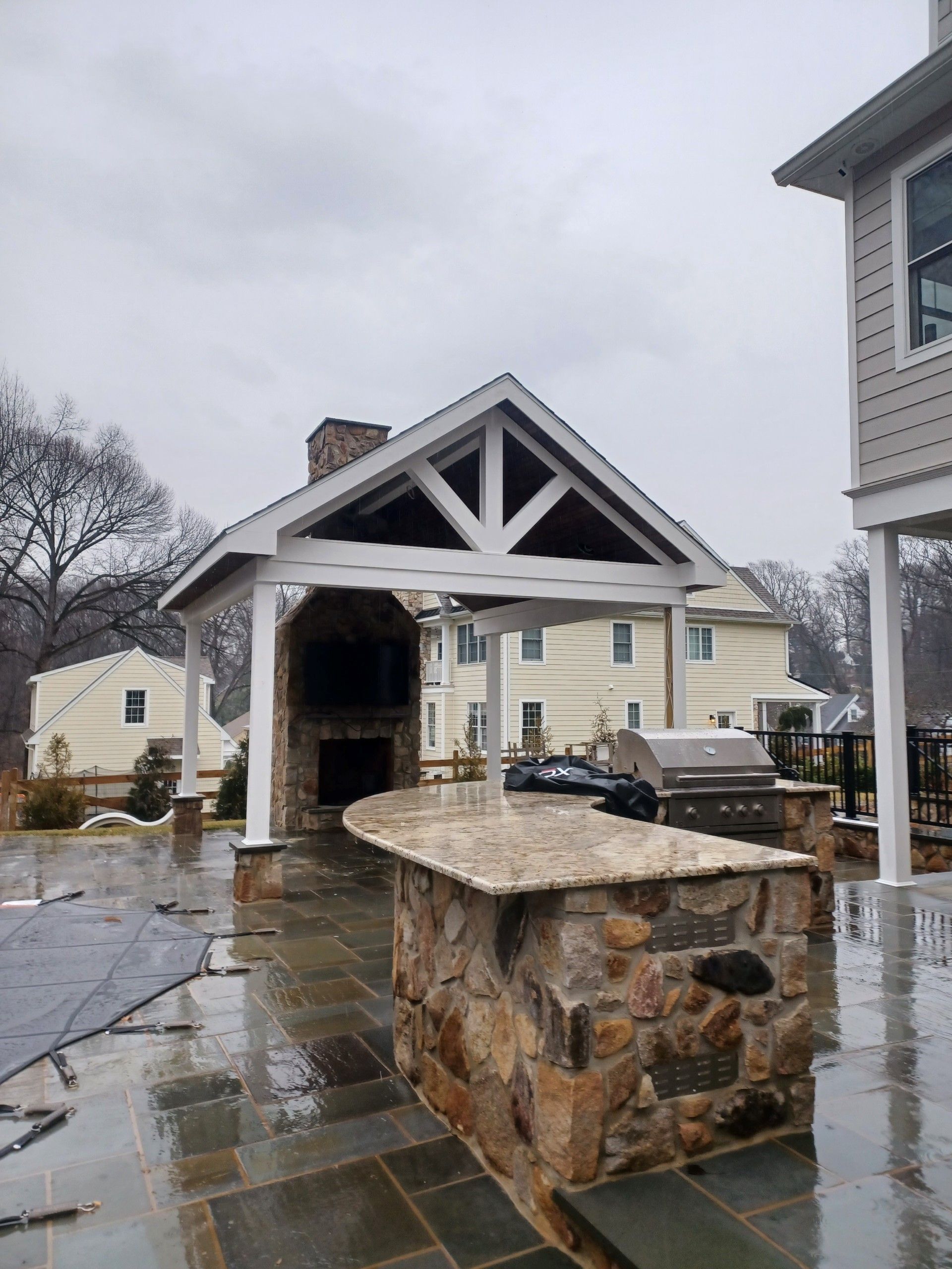 Outdoor kitchen with stone fireplace, bar, and white-trimmed roof under a cloudy sky.