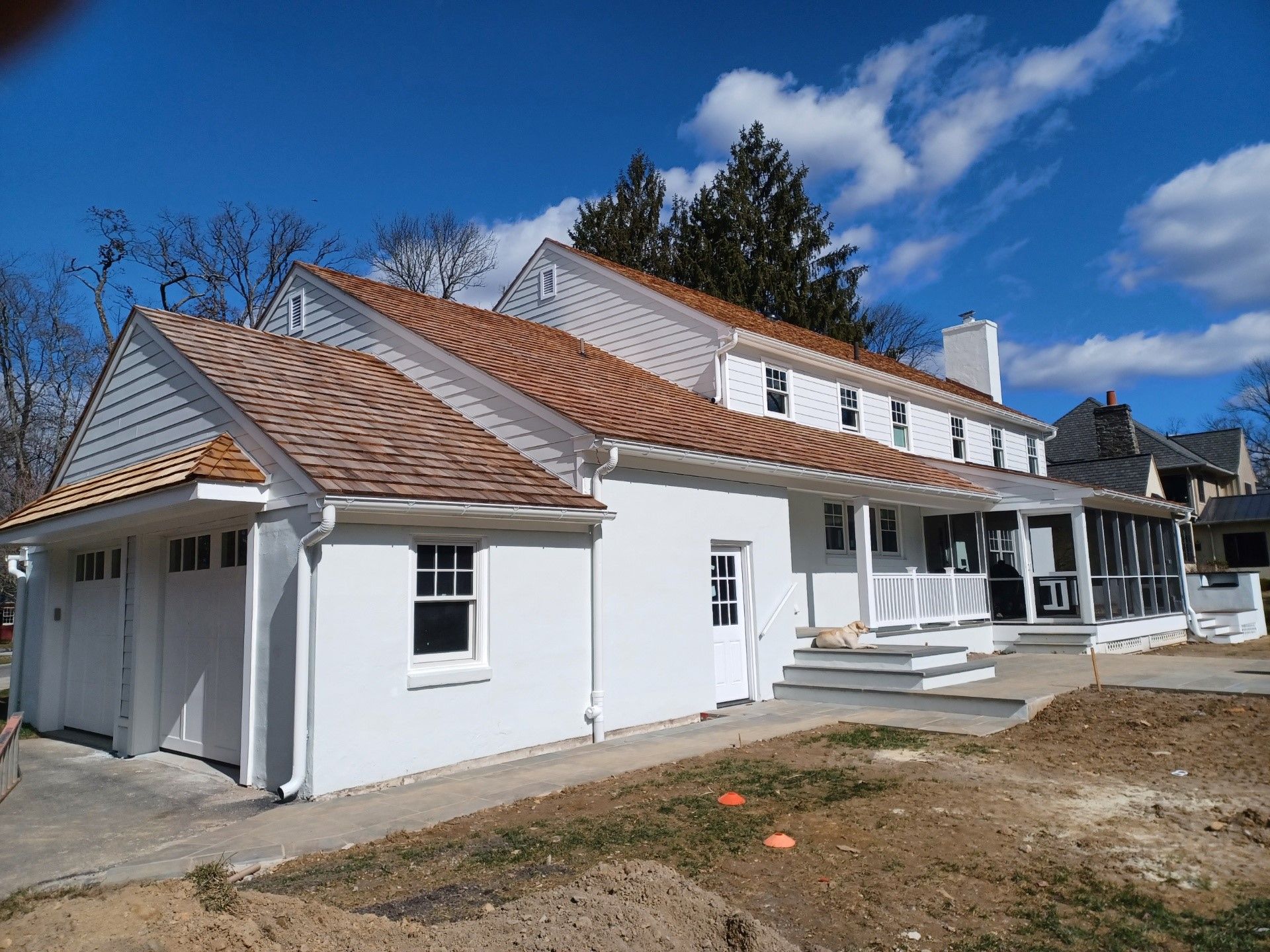White house with a brown shingle roof, garage, and porch on a sunny day.