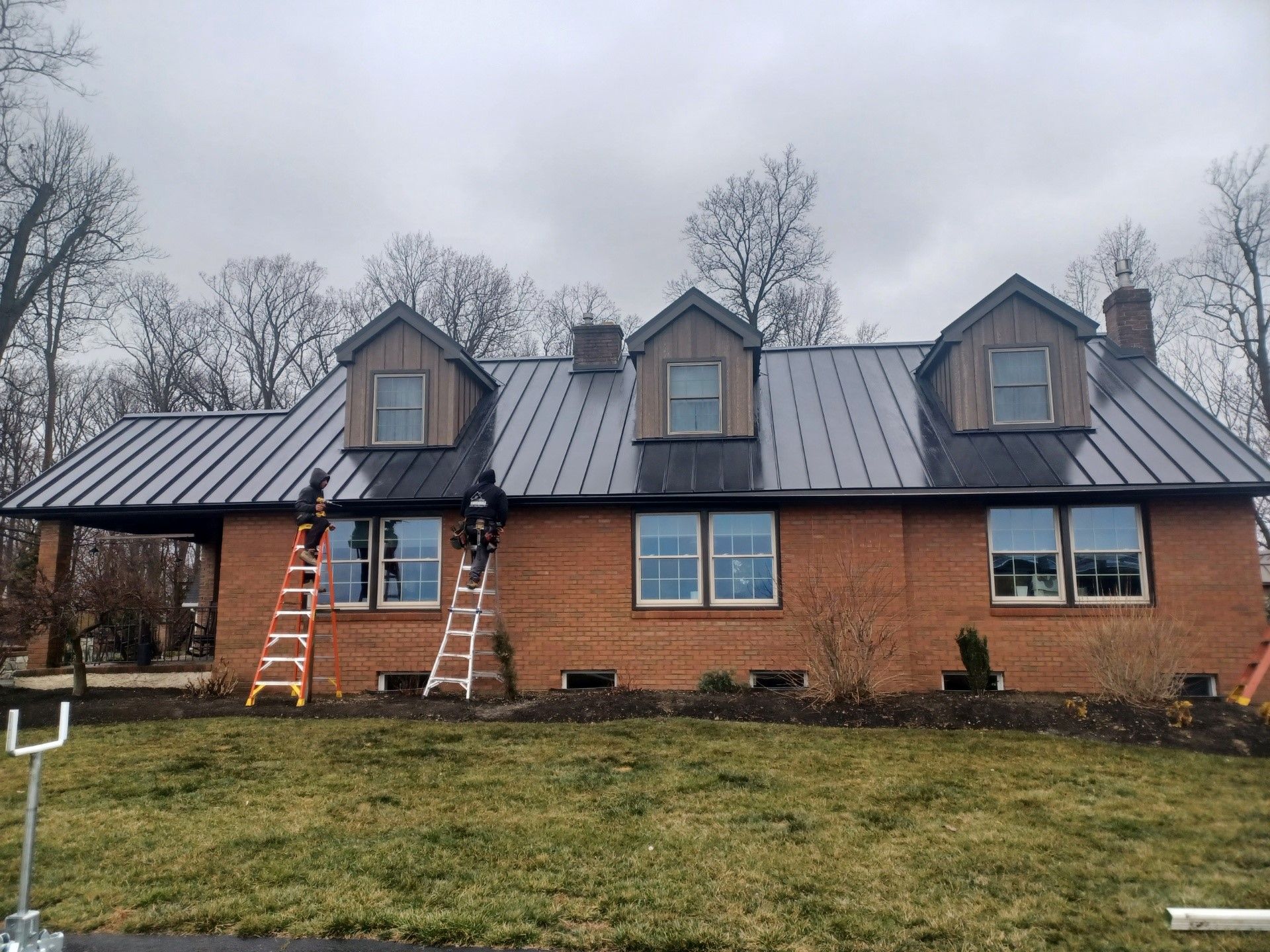 Brick house with black metal roof, three dormers, two workers on ladders, cloudy day.