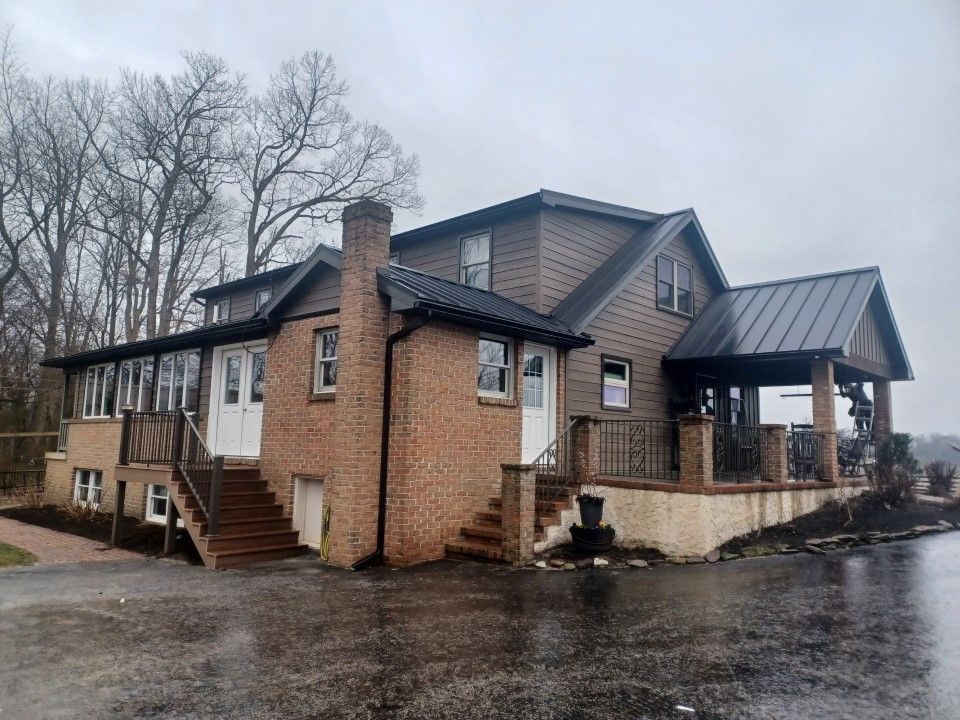 Brown two-story house with black roof and brick accents. Staircase, porch, and bare trees in background.