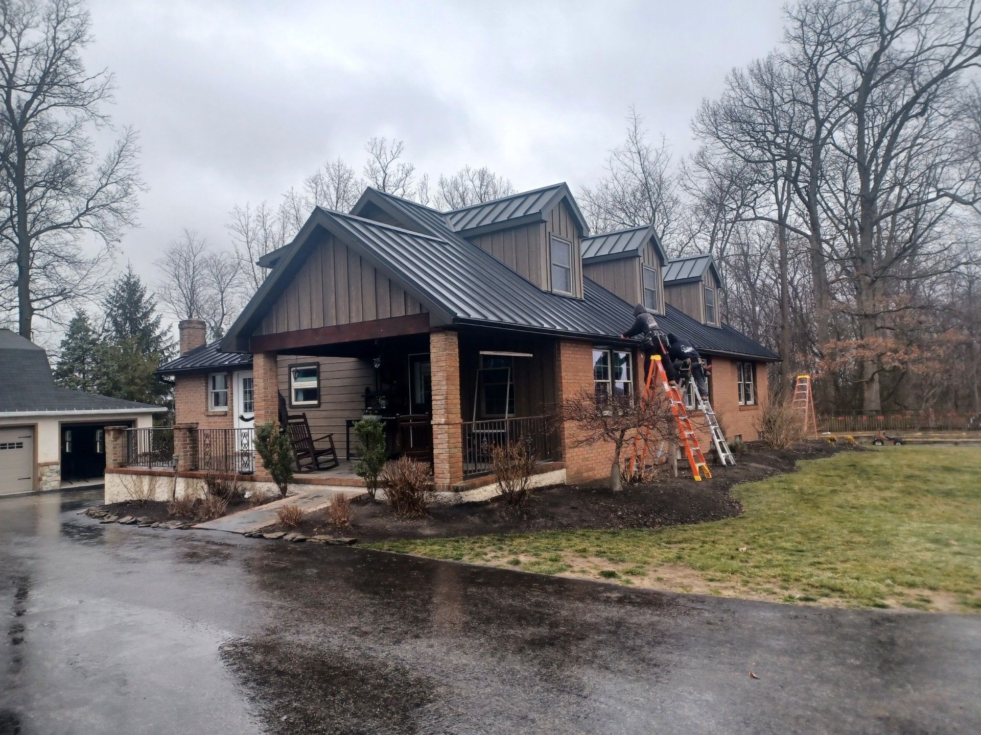 Brick house with dark roof, porch, and brick pillars, parked on a wet driveway, in a tree-filled setting.