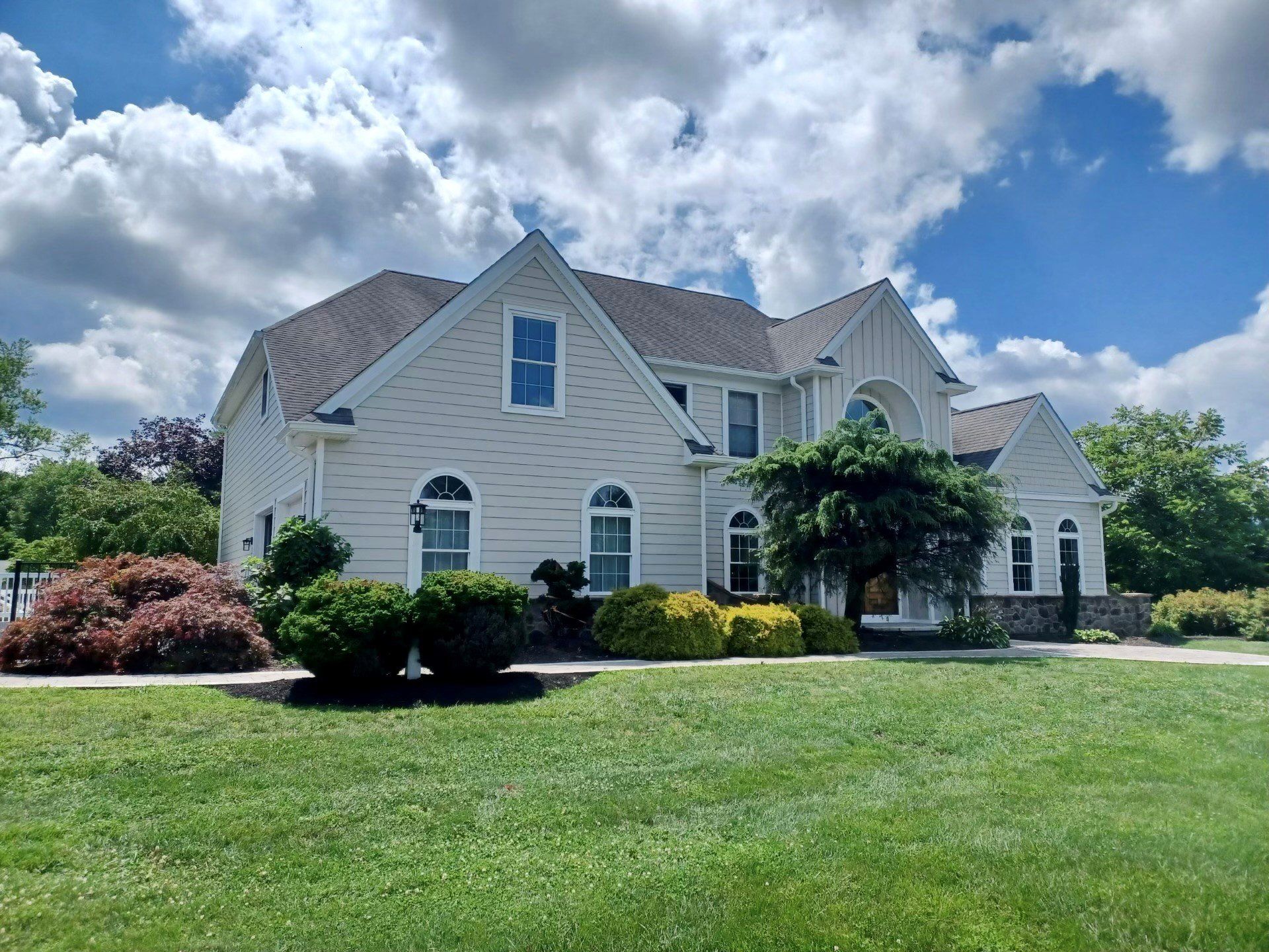 Beige two-story house with arched windows under a cloudy blue sky, surrounded by green grass and shrubs.