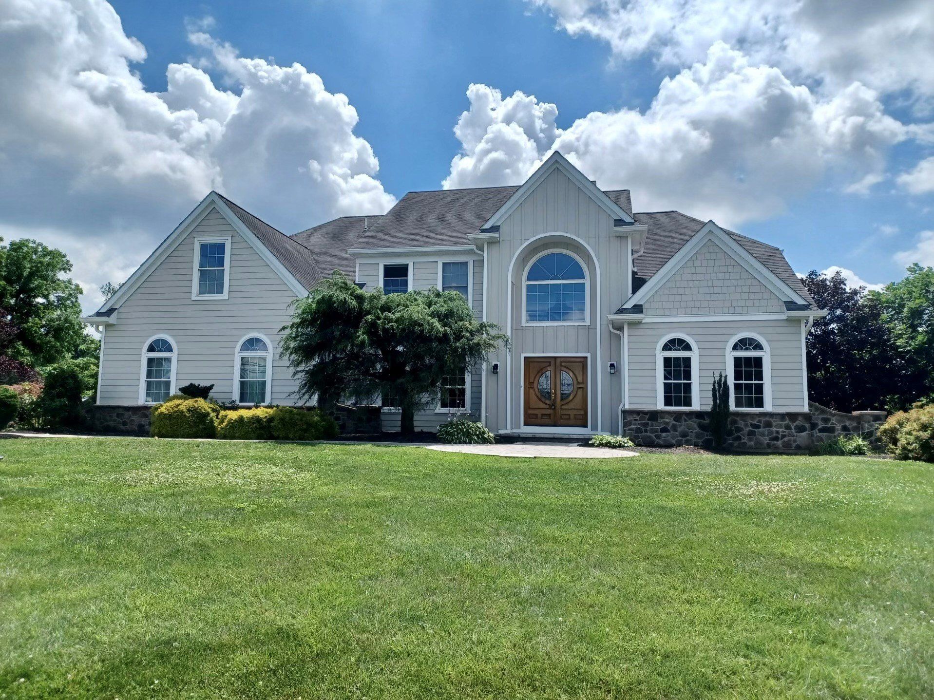 Large, light gray house with arched window, brown doors, and a large lawn under a cloudy sky.