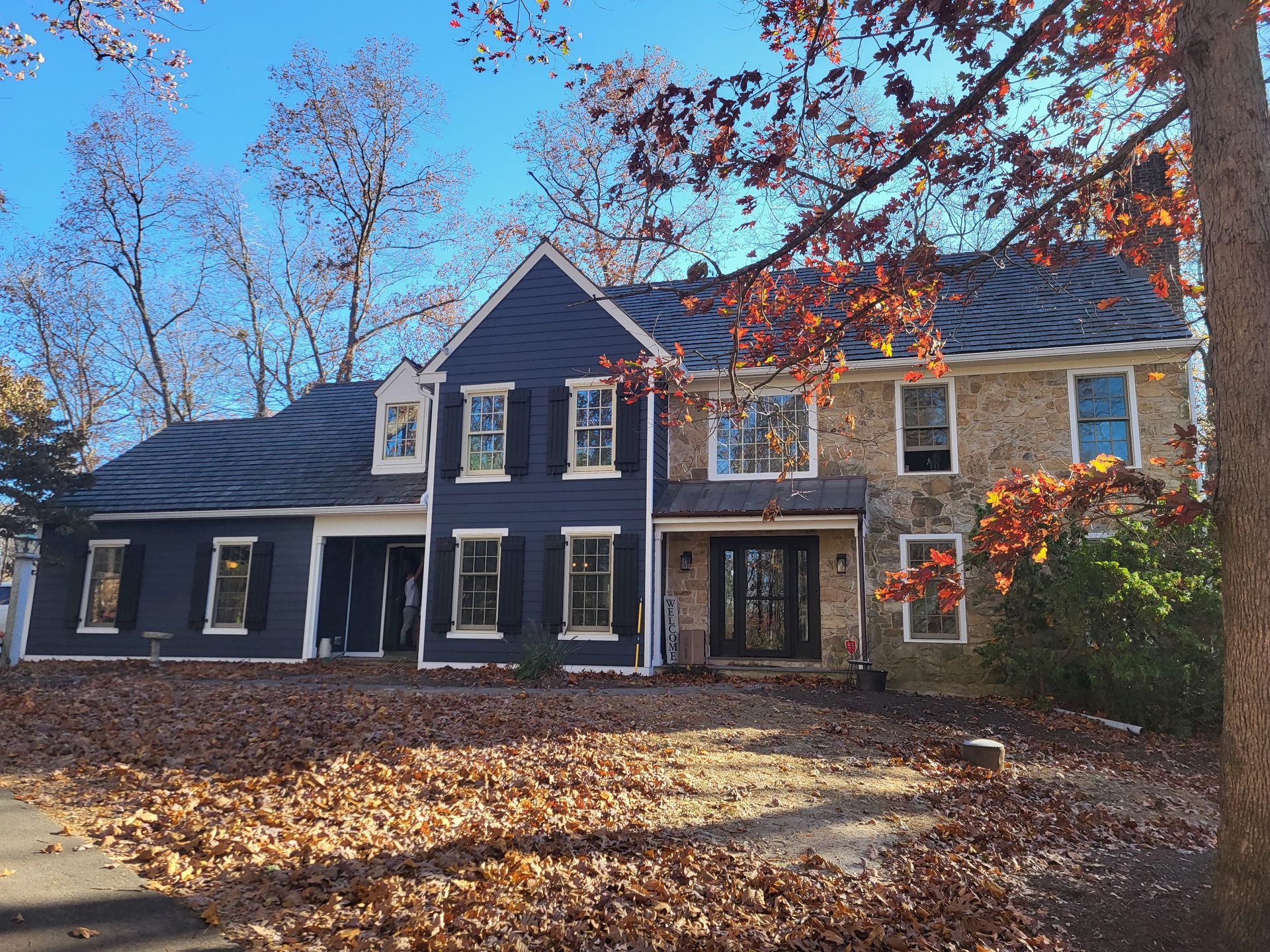Two-story house with blue siding, stone facade, black shutters, and a brown roof. Autumn leaves cover the ground.