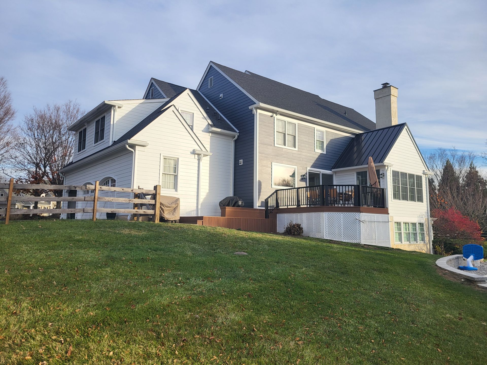 Two-story house with white siding, gray siding, and dark roof on a grassy hill with a deck and fence.