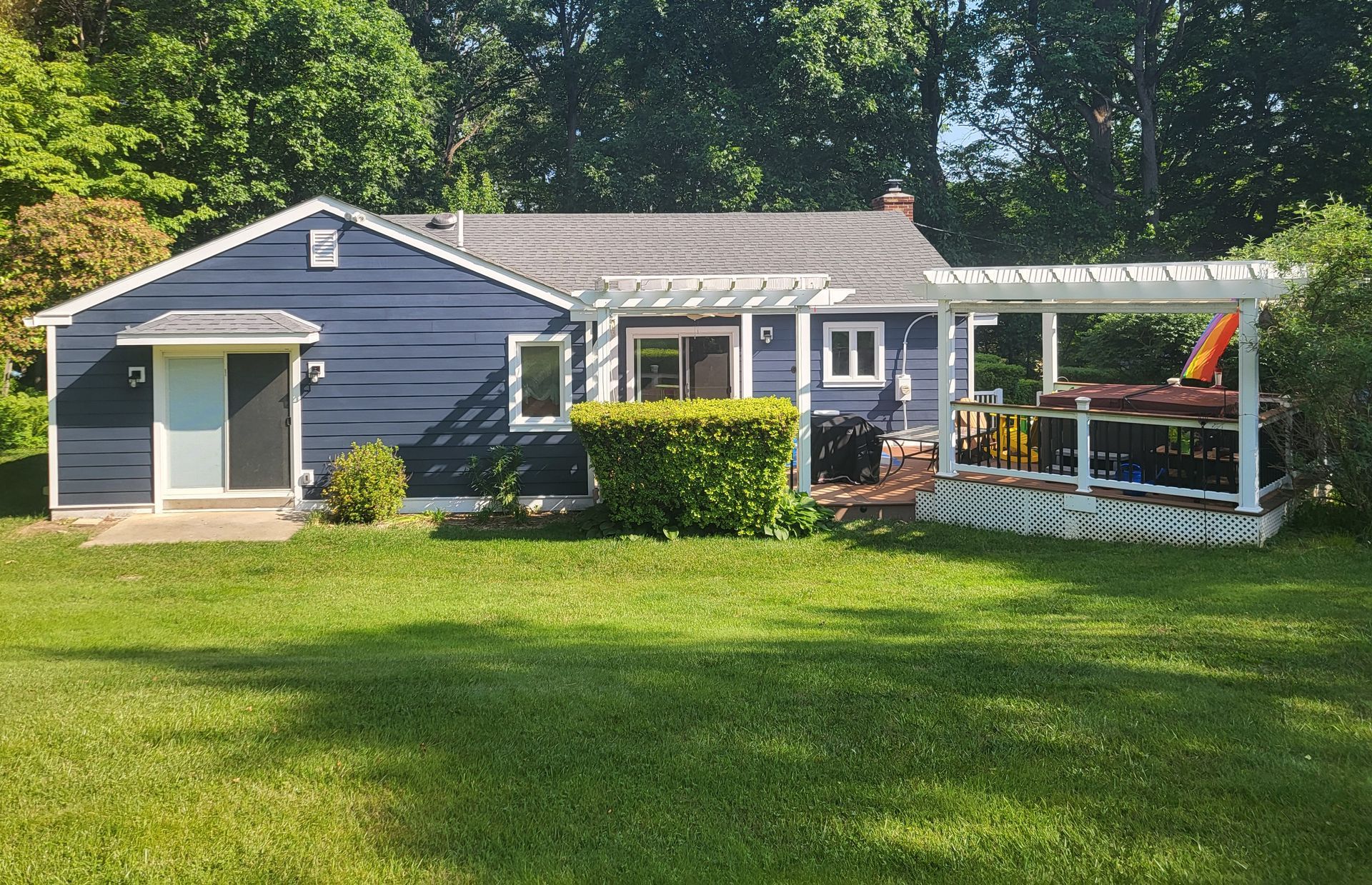 Blue bungalow with white pergola, green lawn, and surrounding trees.