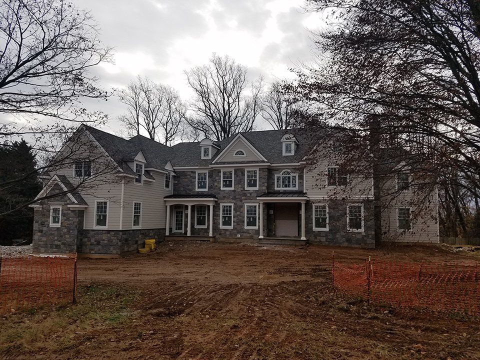 Large, two-story house with gray stone and tan siding. Construction fencing in front; bare trees and cloudy sky overhead.