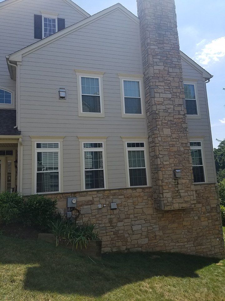 Two-story house with stone chimney, tan siding, and white-framed windows. Green lawn in front.