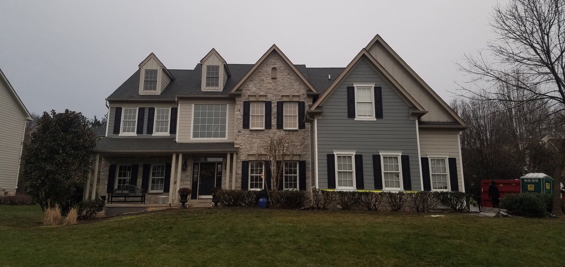 Large two-story house with grey siding, black shutters, and a green lawn under an overcast sky.