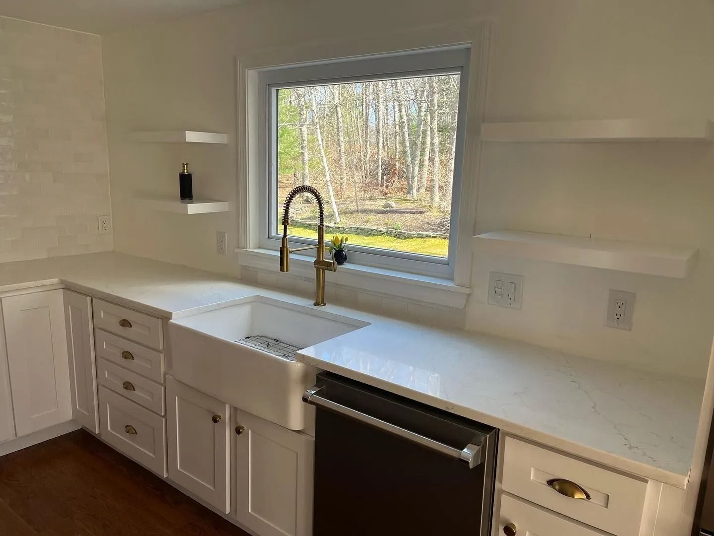White kitchen with a farmhouse sink, gold faucet, and open shelving near a window overlooking trees.