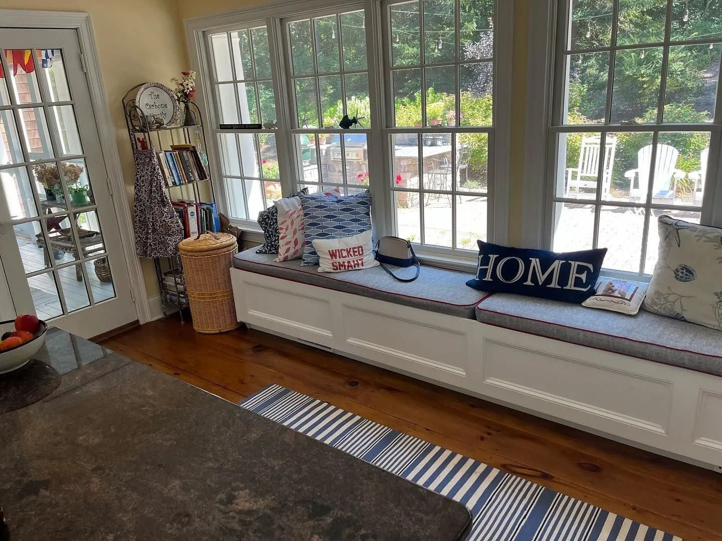 Sunroom with a window seat, cushions, and decorative pillows. Light-filled room with a striped rug and wood floor.