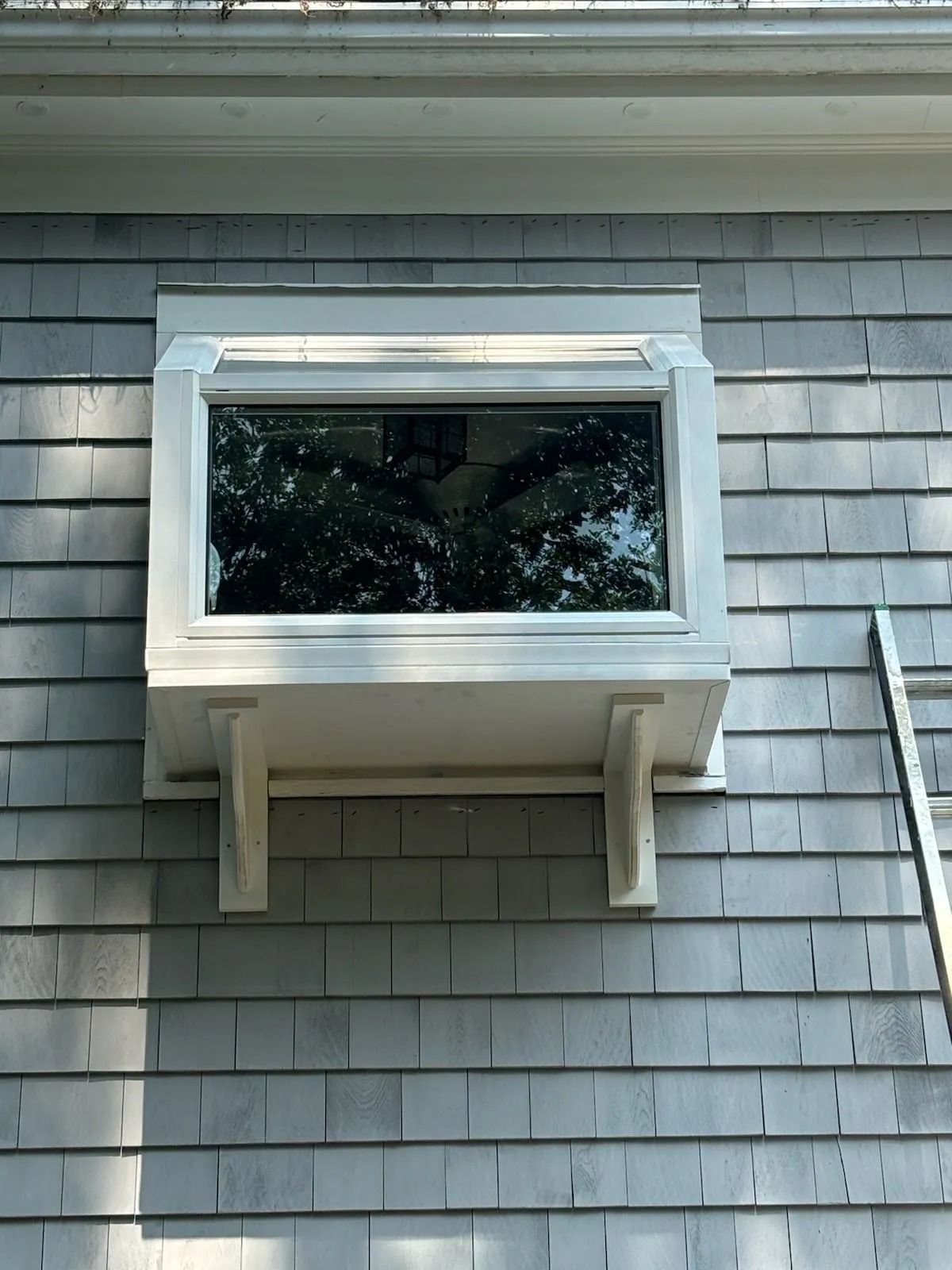 Window box on shingled house exterior. White trim frames the window. A ladder leans against the wall.