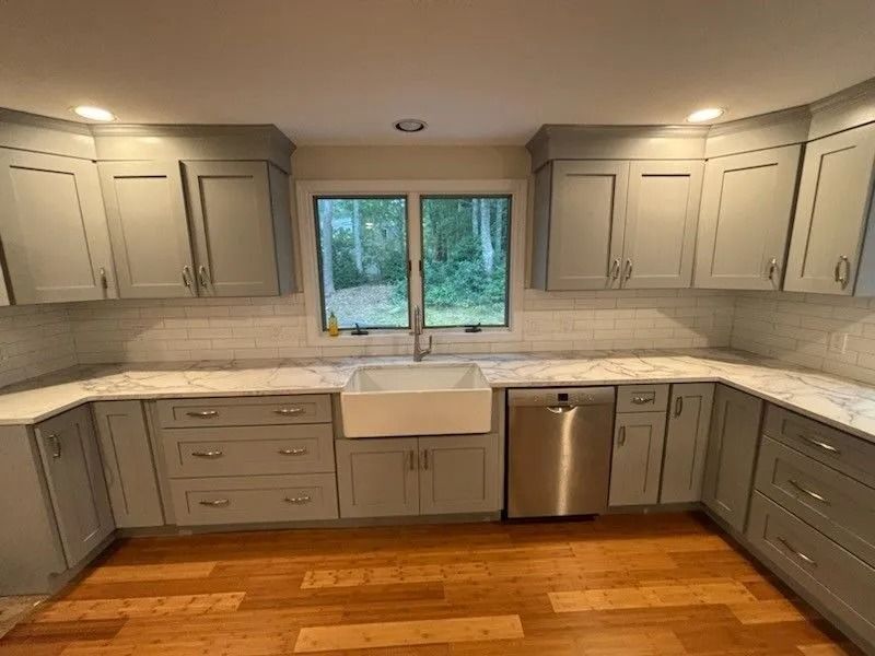 Gray kitchen cabinets with white countertops and a farmhouse sink, stainless steel appliances, and a window overlooking greenery.