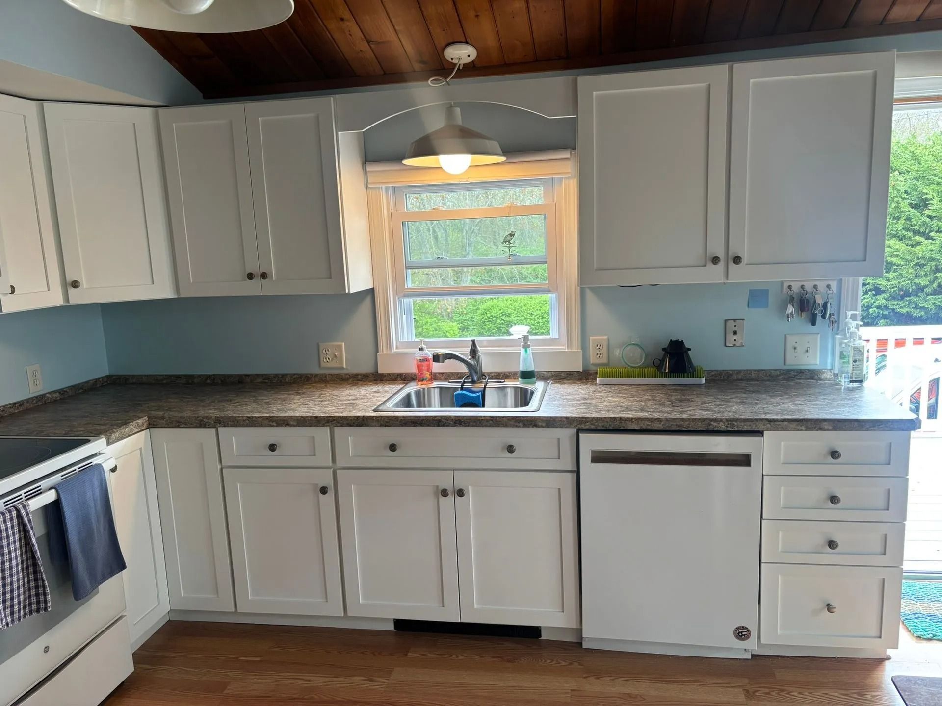 White kitchen cabinets with gray countertops, sink by a window, and a dishwasher.