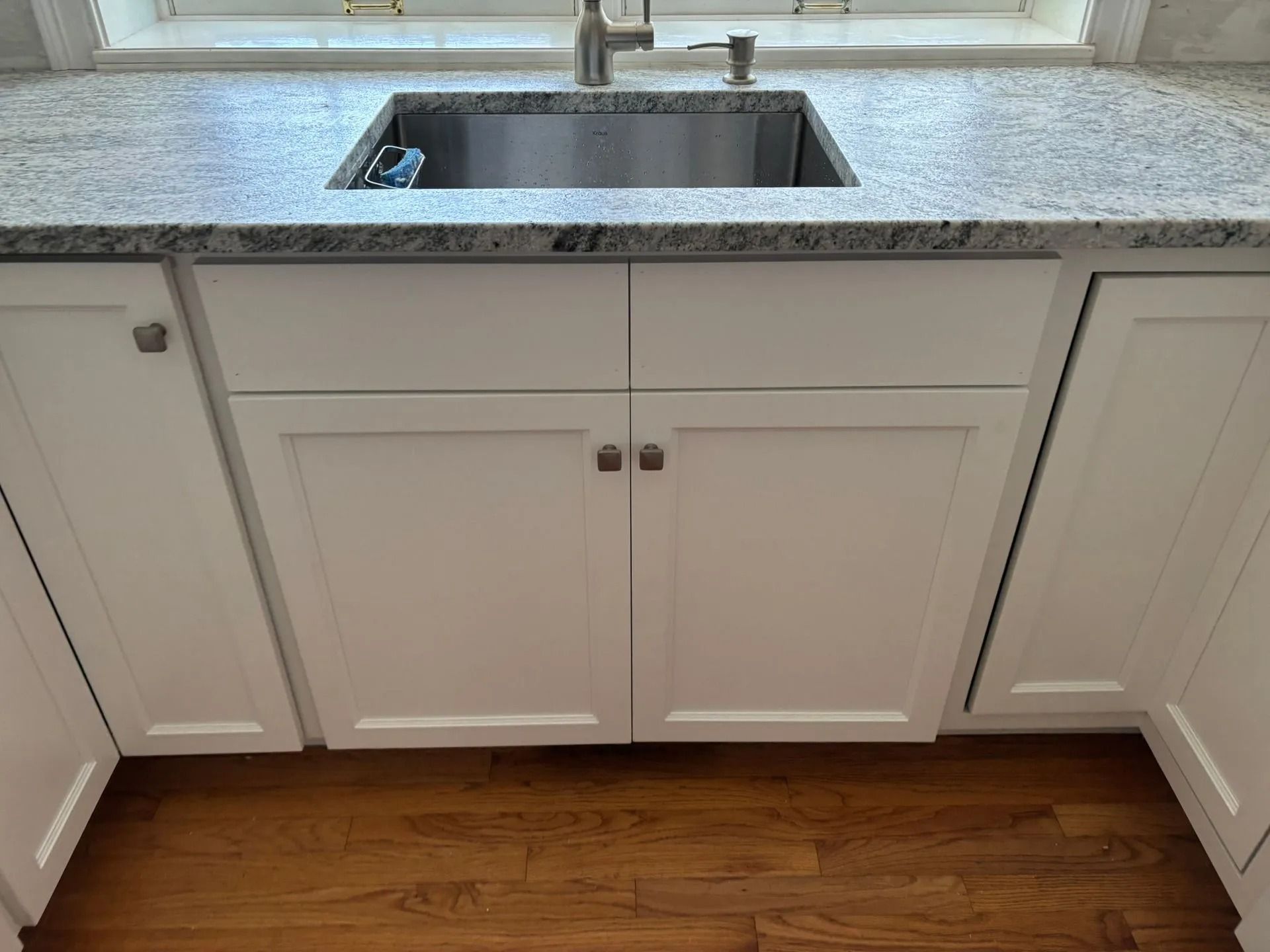 White kitchen cabinets under a stainless steel sink with granite countertop; hardwood floor.