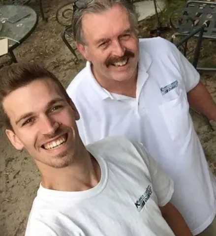 Two smiling men in white shirts, outdoors. One younger, taking a selfie. The other older with a mustache.