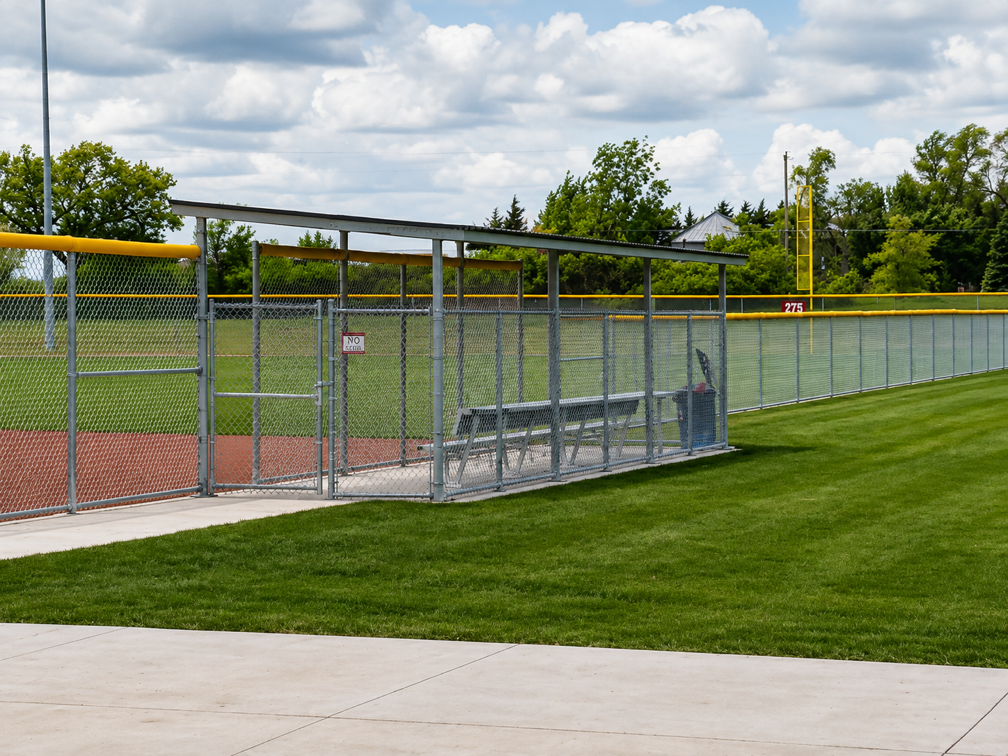 Baseball dugout behind a chain-link fence on a green field under a cloudy sky.