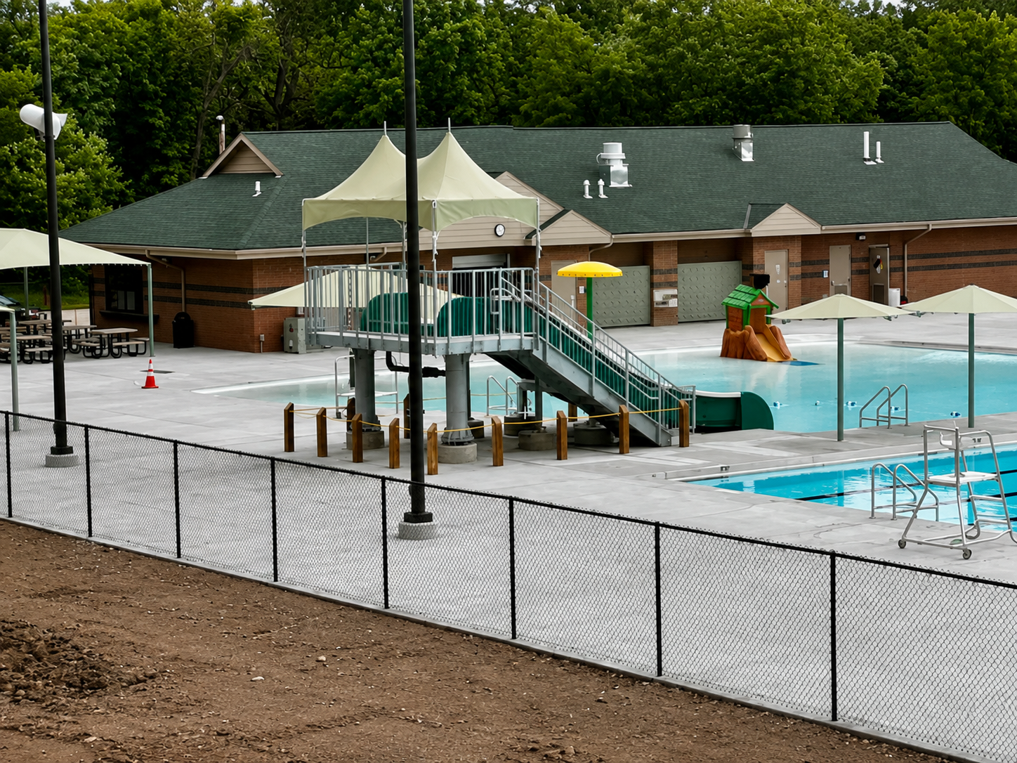 Pool area with slides, fence, and building in the background. Blue water and a light-colored patio surround the pool.