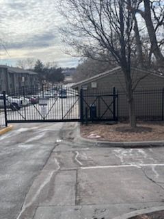 Gated apartment complex entrance with a closed black metal gate. Gray asphalt and a cloudy sky.