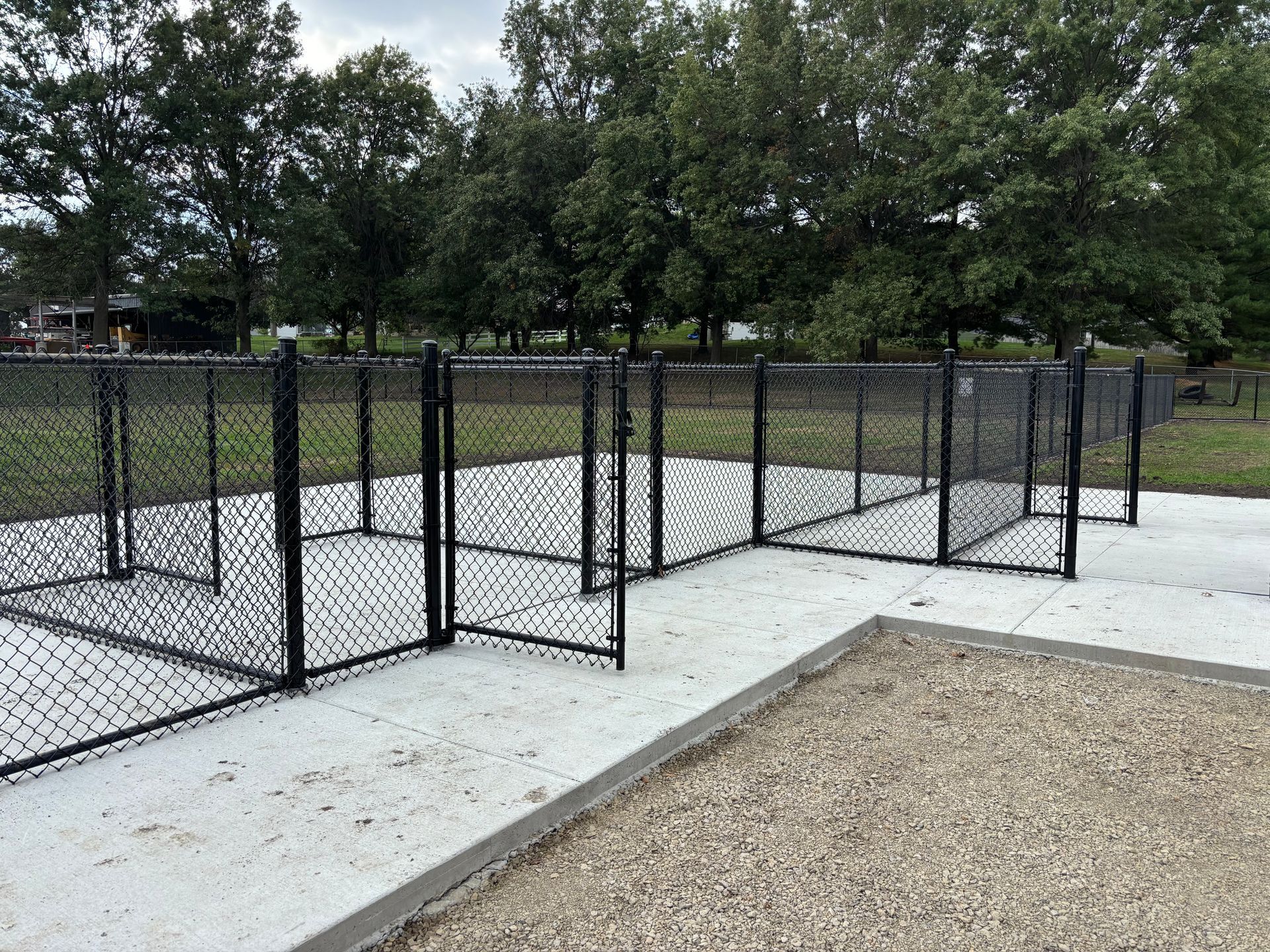 Black chain-link fence dog park with concrete and gravel areas, set against a backdrop of trees.