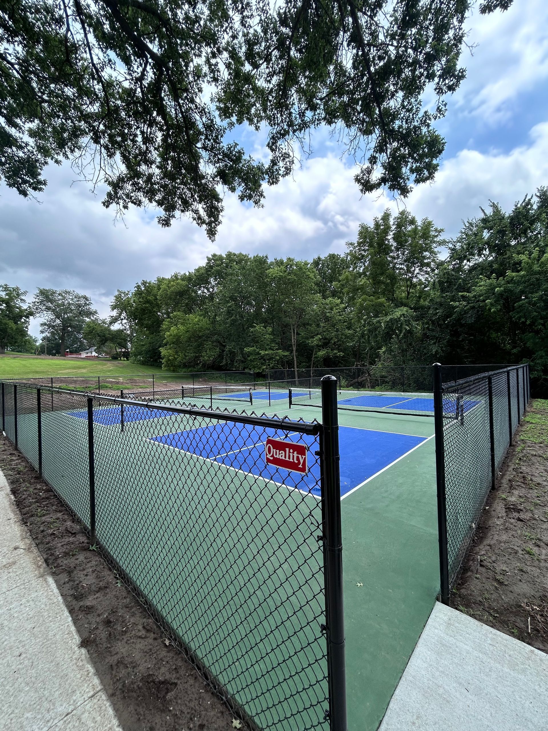 Tennis court with blue and green surface, enclosed by a black chain-link fence, under a cloudy sky.