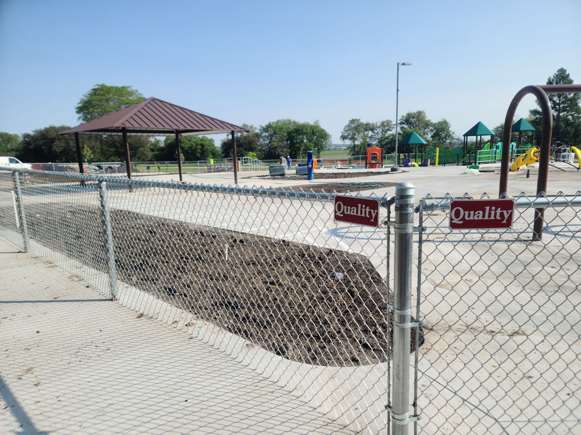 Chain-link fence surrounds a playground under construction, with a shelter and equipment visible.